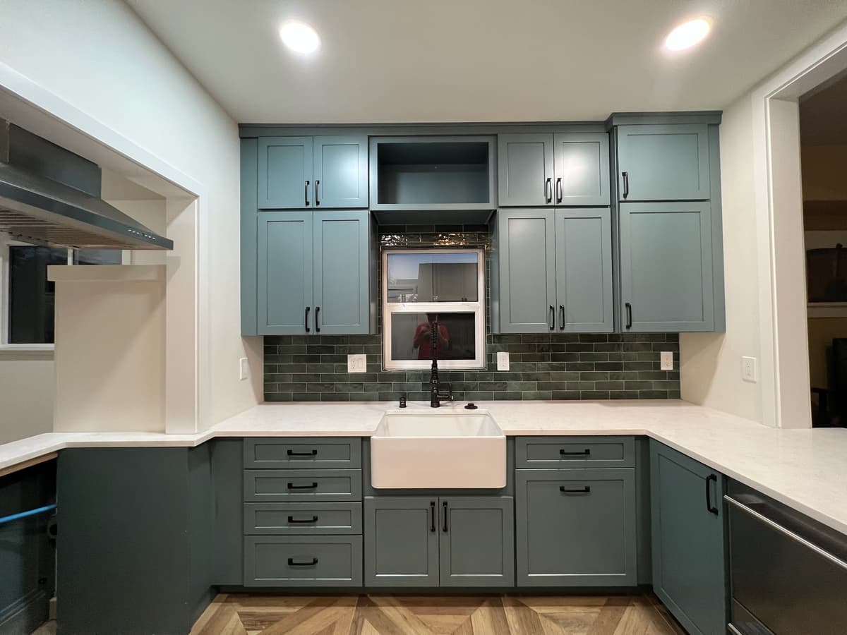 A modern bathroom remodel featuring a double-sink vanity with light wood cabinets and a white countertop. There are three pendant lights above the vanity, a large mirror in the center, an illuminated mirror on the right, and plants in vases for decoration. A door is to the left.